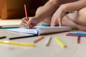 Ground level of unrecognizable kid sitting on floors with legs crossed and drawing in copybook with multicolored pencils