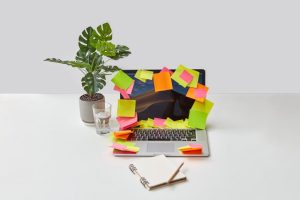Laptop covered with colorful sticky notes on a modern desk with plant and notebook.