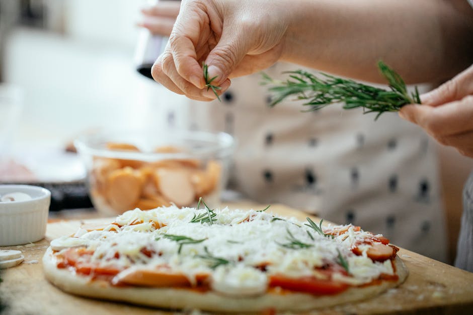 A person adds rosemary to a homemade pizza topped with tomatoes and cheese in a kitchen.
