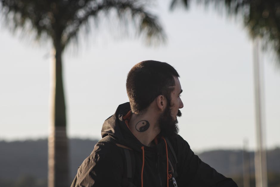 A bearded man with a yin yang tattoo is looking away, framed by palm trees, outdoors.
