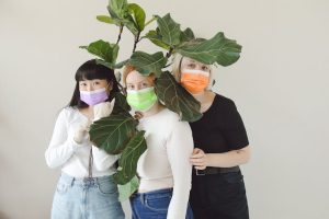 Three women in colorful masks stand behind a fiddle leaf fig, emphasizing pandemic safety and unity.